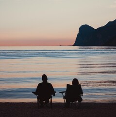 Obraz premium Silhouette of a couple sitting on chairs watching a peaceful sunset over the calm sea, relaxation and mental health concept 