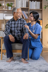 Compassionate Asian female caregiver blue scrubs comforting happy elderly senior man sitting chair...