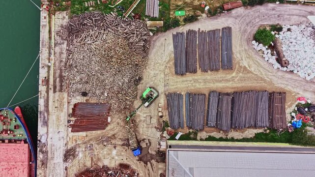 Machinery moves around the construction site as workers organize metal and wood materials on the ground next to the river. The scene shows ongoing building activities.