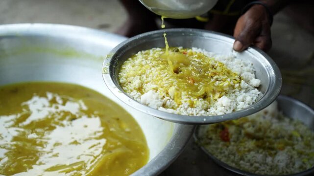 Natore, Bangladesh - 18 May 2024: View of a person pouring yellow dal over white rice in a metal plate from a large communal bowl.