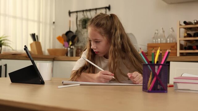 A young girl sitting at a kitchen table, drawing or writing on a notebook while looking at a tablet propped up nearby.