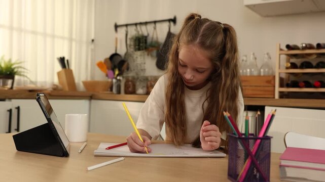 Young girl with long hair drawing or writing with colored pencils at a kitchen table, focused and engaged in creative activity.