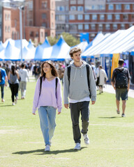 Obraz premium Two young people walk through an outdoor event with white tents and a crowd in an urban setting on a sunny day.