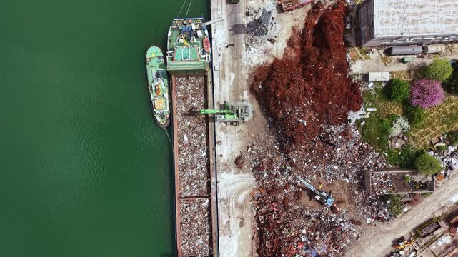 A barge is docked near a recycling facility with piles of waste on the shore. Workers operate machinery to sort and remove materials.