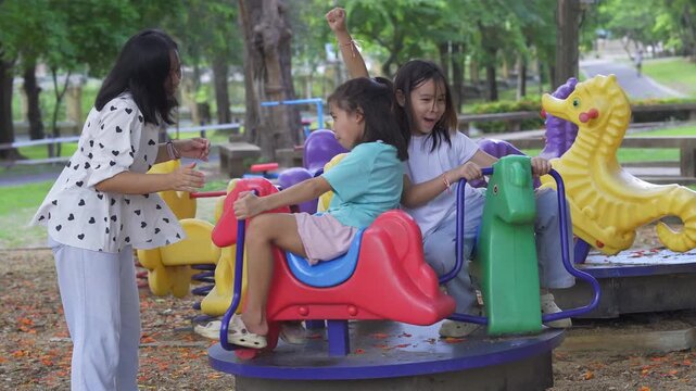 Happy Asian mother pushing her two young daughters on a colorful playground roundabout merry-go-round.