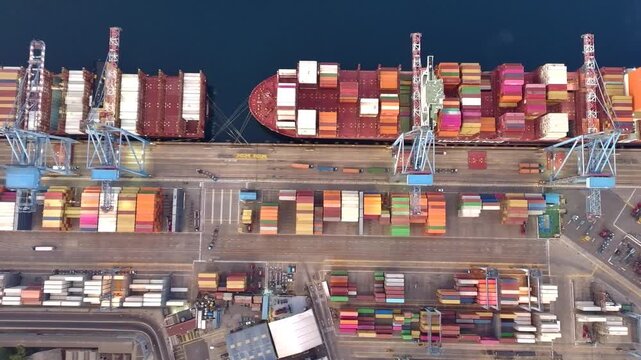 Aerial top view of a container cargo ship docked at a busy commercial port terminal with colorful shipping containers, cranes and logistics infrastructure at dusk.