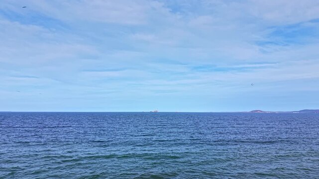 Waves crash gently against the beach as boats sail in the distance. The sky is clear and blue with a few clouds, providing a bright setting for the scene.