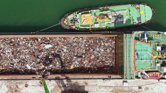 A cargo vessel is loading scrap materials at the port with a tugboat helping to secure the ship. The scene captures the busy activity during daylight hours.