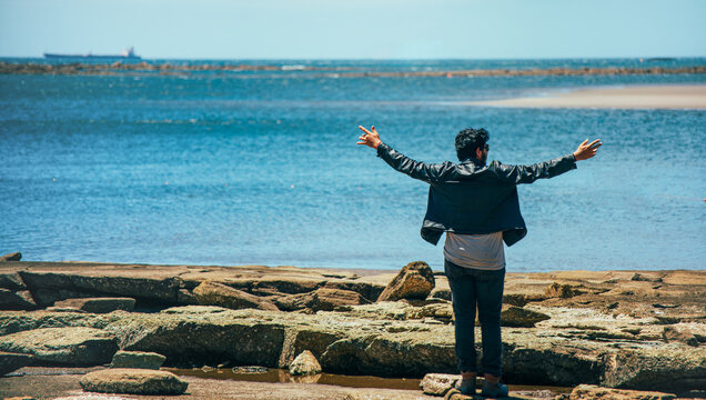Man standing on the rocky shore with his arms outstretched looking out at the ocean. Man with his back to the camera, arms outstretched, looking out to sea