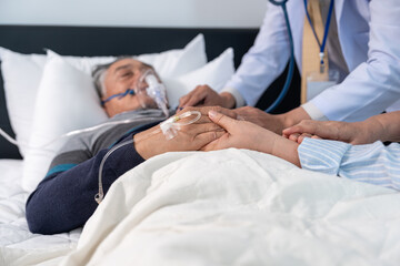 Obraz premium scene of senior woman holding hands an elderly male patient dying on bed that on iv drip and oxygen mask while a doctor using stethoscope examine on chest, selective focus at hands