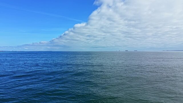 A vast expanse of ocean stretches out under a mostly clear sky. Soft clouds drift above with a few ships visible on the horizon far away from the shore.