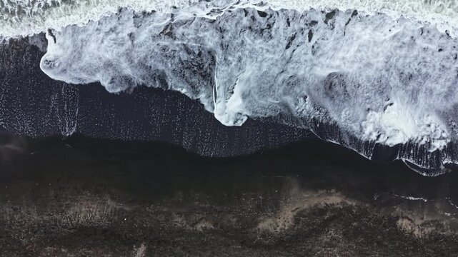 Waves crash onto a dark sandy beach under bright daylight. The white foam contrasts against the dark sand while patterns form in the water.
