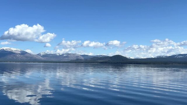 4K HD Video traveling across the water from forested river bank to snow capped mountains along the shoreline of Lake Tahoe. Clouds reflected in the crystal blue calm water.
