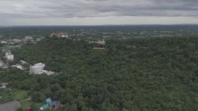 Aerial drone view of Sakae Krang Hill in Uthai Thani, Thailand, featuring the golden "City of the Chakri Dynasty's Progenitor" sign and hilltop temple against a cloudy sky and lush green forest.