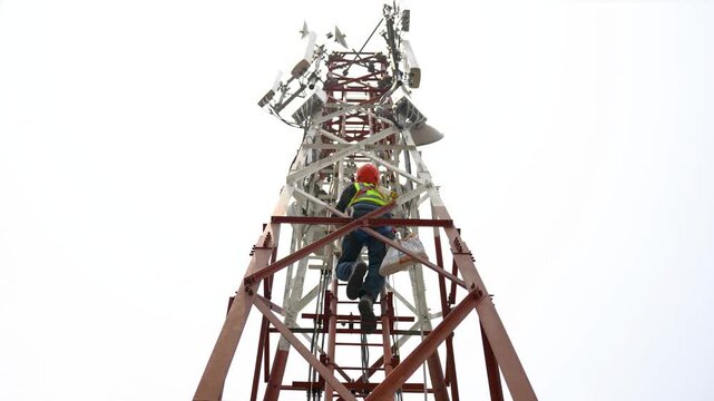 Dhaka, Bangladesh - 31 January 2024: View of a technician in a safety vest climbing a tall telecommunications tower against a bright sky.