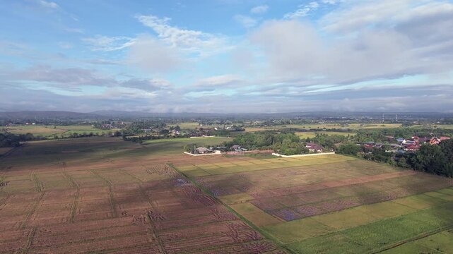 A cinematic aerial drone view capturing a vast agricultural valley characterized by diverse crop patterns and field textures, transitioning into a lush green landscape featuring a peaceful rural villa