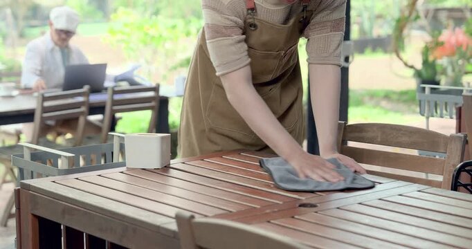 Asian son wipes wooden cafe table with cloth near tissue box while father works behind, practicing hygiene checklist, service readiness, family restaurant training, detailed maintenance duty and care.