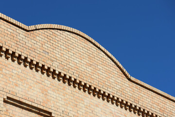 An architectural detail of a light tan brick building facade featuring a curved parapet wall and decorative corbeled brickwork against a clear blue sky © Tammy Walker