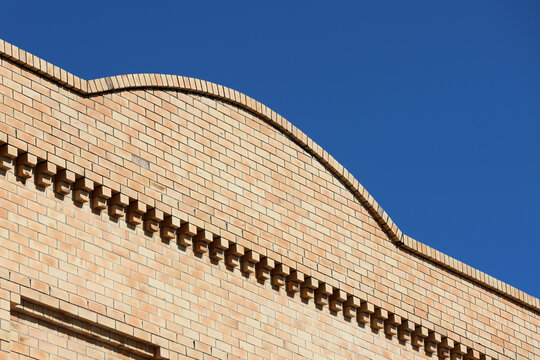 An architectural detail of a light tan brick building facade featuring a curved parapet wall and decorative corbeled brickwork against a clear blue sky