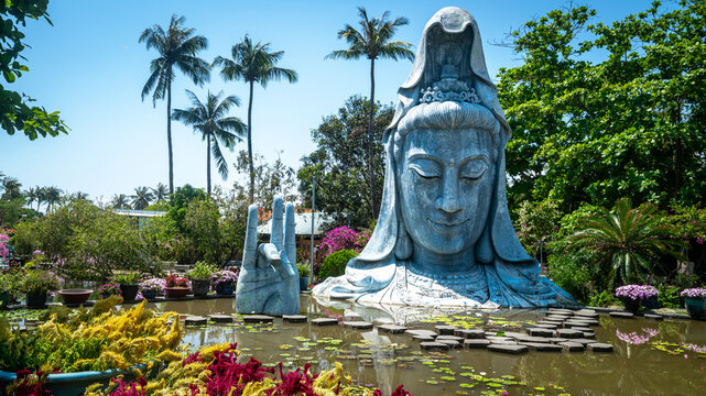 Large white Quan Am (Guanyin) Buddha statue at Chua Thanh Luong Buddhist Temple, Phu Yen Province, Vietnam. The serene bodhisattva figure is surrounded by tropical palm trees and colorful flowers in a