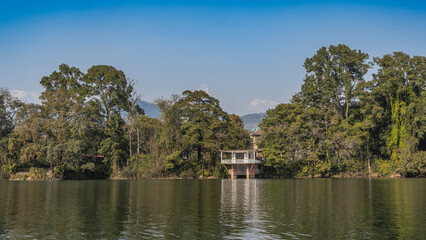Fototapeta premium A beautiful lake. On the shore, among the green trees, there is a gazebo with viewing platforms. Himalayan mountains in the distance against a clear blue sky. Nepal. Pokhara. Lake Phewa.