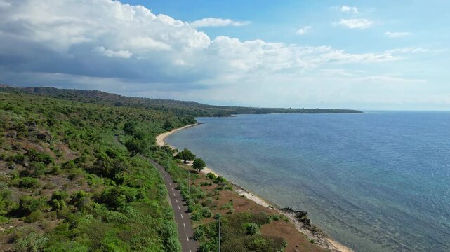Aerial drone footage of a long coastal road in Soromandi area, Bima regency, Sumbawa island, Indonesia, with blue sea waters and reefs, white sand beach, near Rambu Suar Batu Besar lighthouse