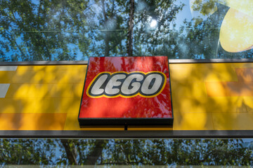 Obraz premium Germany, Berlin May 23, 2026 A LEGO store sign stands against a yellow facade and reflective glass. Reflections of London plane trees and blue sky add color to the urban storefront.