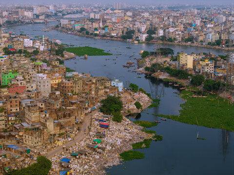 Aerial view of the Buriganga River flowing through densely packed urban buildings with small boats and green aquatic plants Keraniganj, Dhaka Division, Bangladesh.