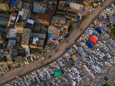 Aerial view of a busy street and dense urban buildings next to a large waste sorting area with white sacks and colorful tarps in Keraniganj, Dhaka Division, Bangladesh.