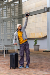 Adult male traveler in beanie, denim jacket, headphones, holding suitcase, raising phone in plaza © wavebreak3