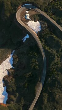 Vertical Screen: Beartooth Highway in Montana USA captured by drone showing serpentine mountain road with melting snow patches and green alpine vegetation during golden hour light.