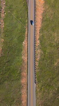 Vertical Screen: Beartooth Highway in Montana USA as a white car travels along the winding road with yellow center line and metal guardrail through lush green grasslands.