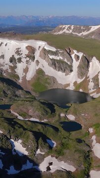 Vertical Screen: Beartooth Highway Montana USA drone footage reveals stunning alpine lakes nestled in glacial cirques with snow patches dotting green meadows and rugged mountain peaks under clear.