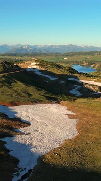 Vertical Screen: Beartooth Highway in Montana USA winds through alpine terrain with melting snow patches and a pristine blue lake surrounded by lush green meadows and distant peaks.