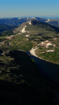 Vertical Screen: Beartooth Highway in Montana USA features stunning alpine landscape with mountain lake and scattered snow patches across green meadows under clear blue sky.