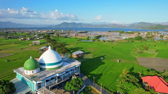 Aerial drone footage of Darul Hamid islamic center, a vast salt pond field valley nearby the sea, in Bolo area, Sumbawa, Indonesia, and green rice fields aside, mountains behind with Bima city bay