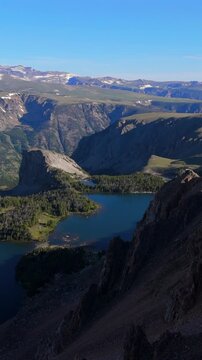 Vertical Screen: Beartooth Highway Montana USA aerial drone view of stunning alpine lake nestled in deep mountain canyon with snow-capped peaks and evergreen forests on clear summer day.