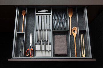 Obraz premium Modern kitchen drawer organizer with neatly arranged cooking utensils, knives, scissors, wooden spoons, and kitchen accessories in minimalist dark interior design. Clean and organized storage concept 