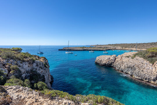 Multiple sailboats and motorboats moored in stunning crystal clear turquoise waters of secluded Cala Varques bay on a sunny summer day in Mallorca, Balearic Islands