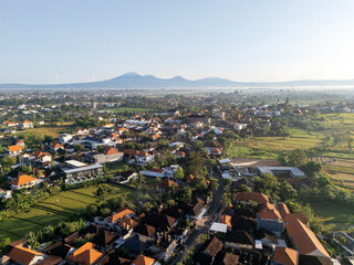 Obraz premium Aerial panoramic view of a lush Balinese village in Canggu with terracotta roofs and distant mountains under a warm golden sunrise.