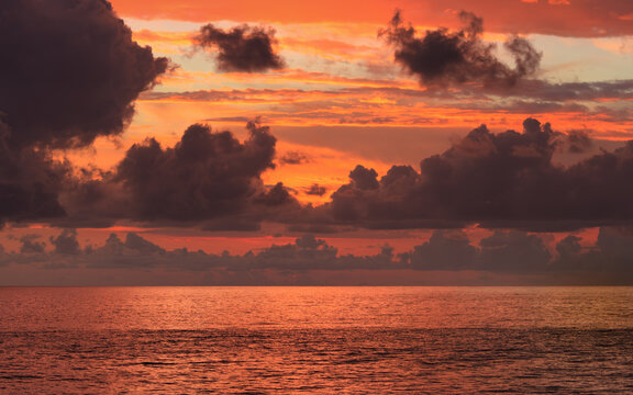 Fiery sunset sky above calm ocean horizon with dark clouds and orange reflections on water surface. Tropical sunset, atmospheric seascape with vivid twilight colors. Weather forecast concept
