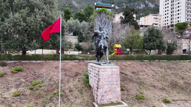 Aerial tracking shot circling the equestrian statue of national hero Gjergj Kastrioti Skanderbeg next to a waving Albanian flag, with the mountainous backdrop and buildings of Kruje