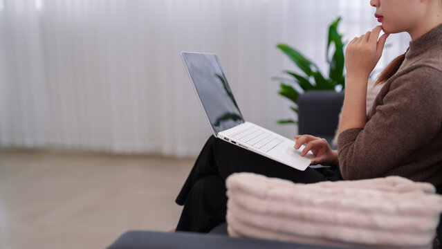 Asian businesswoman working from home using laptop computer sitting on couch thinking planning strategy business idea remote work office space plant
