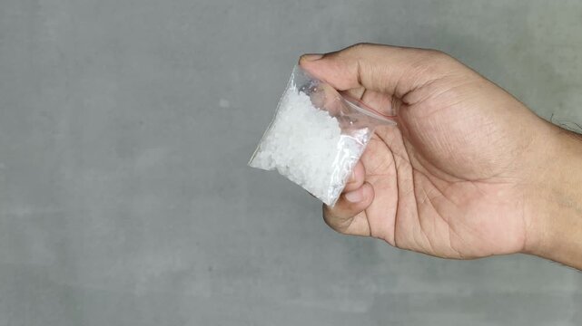 Closeup of asian or indian person hand holding lab made synthetic drug methamphetamine or crystal meth inside small zip bag isolated on white background. Consuming illegal narcotic substance concept.