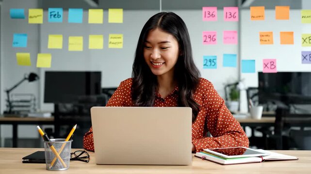 Smiling young Asian businesswoman working on laptop in modern office with sticky notes on wall.