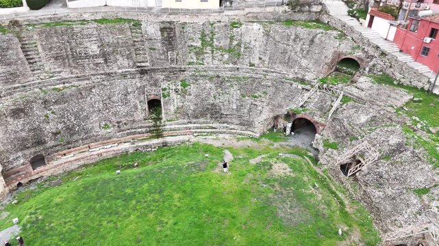 High-angle aerial drone shot panning right across the curved stone seating rows, green grassy arena, and ancient brick tunnel entryways of the historic Roman Durres Amphitheatre in Albania