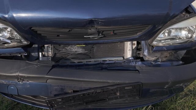 Damaged front of a dark blue car after a collision. Slow motion broken headlights, torn bumper, dented grille, wrinkled car bonnet and missing license plate.