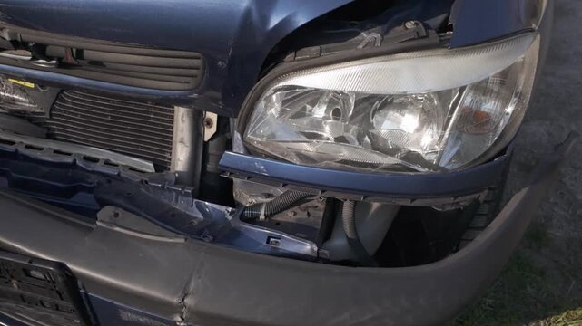 Damaged front of a dark blue car after a collision. Slow motion dolly out close-up of left headlight, torn bumper, dented grille, wrinkled car bonnet and missing license plate.