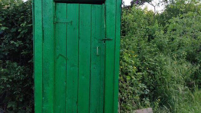 Person walking towards rustic wooden outhouse, opening latch and revealing toilet hole inside, first person view