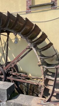 Antique Water Wheel Mechanism: A close-up view of an old, rusty water wheel, its wooden paddles worn by time and use, set against a textured wall. Robbiano, Italy